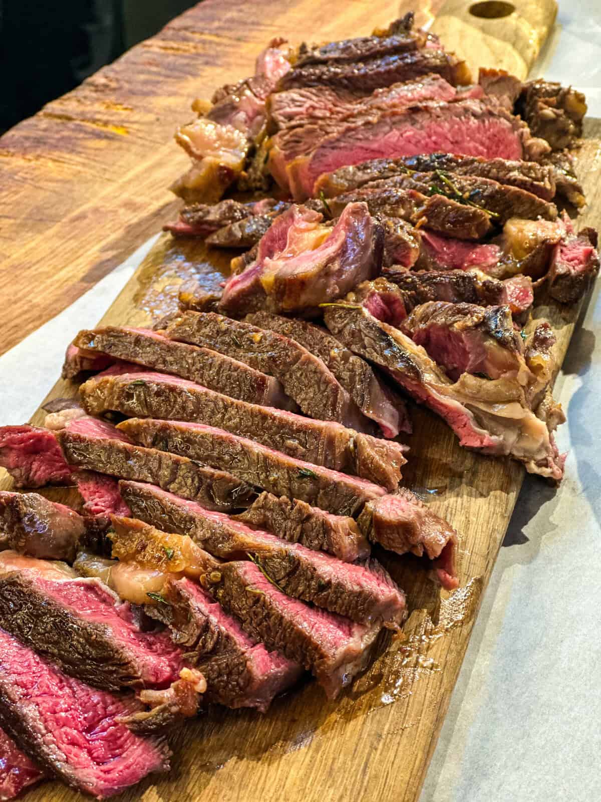 Sliced medium-rare steak resting on a wooden cutting board, showing a juicy, pink center and seared brown crust. The steak pieces are arranged in overlapping rows.