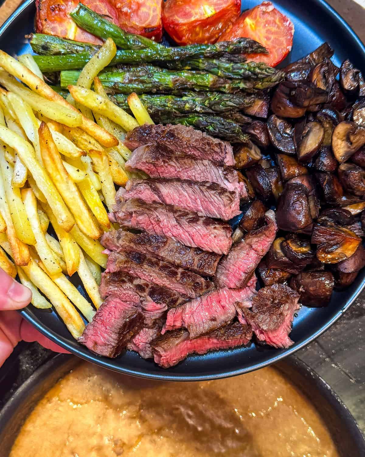 A plate with sliced medium-rare steak, roasted mushrooms, asparagus, tomato halves, and golden French fries is held above a pot of simmering brown gravy.