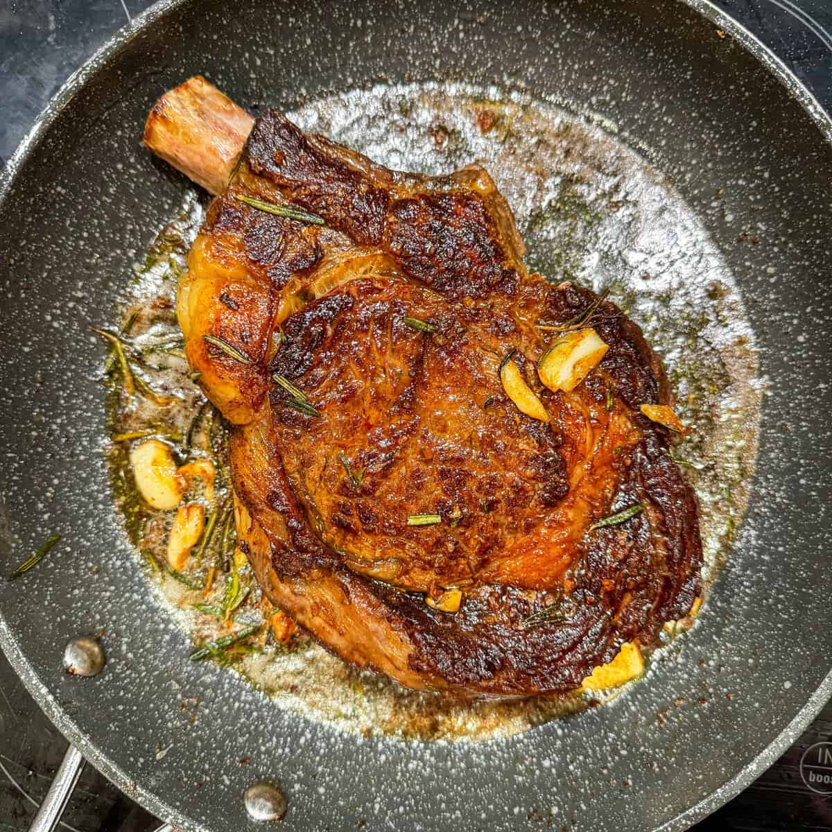 A seared ribeye steak with a browned crust cooks in a skillet, surrounded by melted butter, sliced garlic, and sprigs of rosemary.