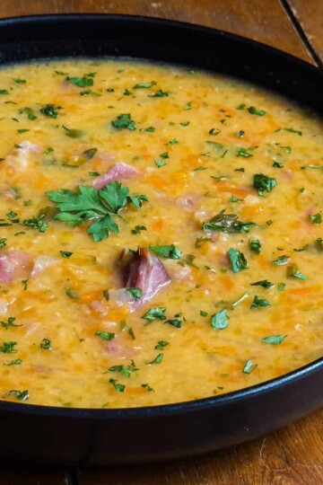 A black bowl filled with a thick, scottish lentil and ham soup is placed on a wooden table. The soup is garnished with chopped parsley and contains visible pieces of meat and vegetables.