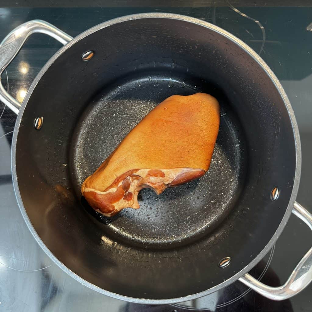 A single smoked ham hock is resting at the bottom of a large, empty black pot with two handles, placed on a stovetop. The ham hock has a rich brown color, and the pot has a shiny, slightly reflective surface.