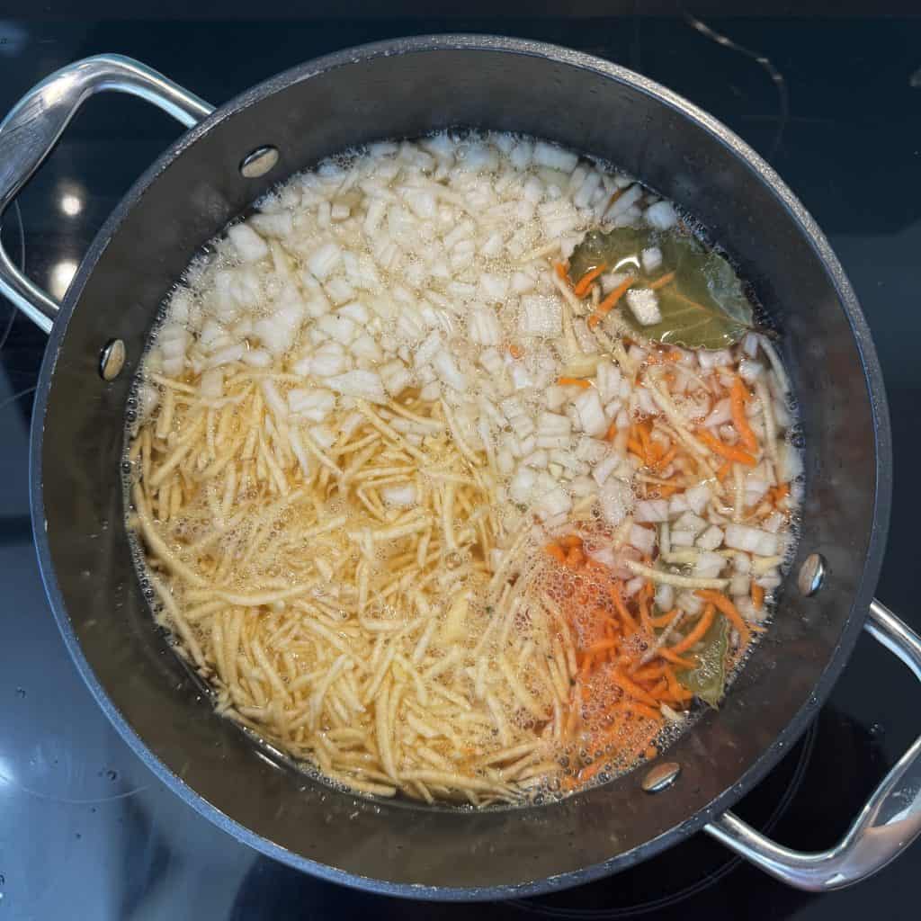 A pot on a stove contains boiling soup with chopped onions, shredded carrots, and bay leaves. Thin noodles are also visible, partially submerged in the bubbling broth. Two metal handles are on the sides of the pot.