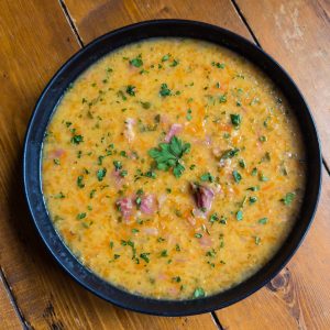 A black bowl filled with a hearty soup on a wooden table. The soup has a creamy texture with visible pieces of meat and is garnished with chopped parsley and a parsley leaf in the center.