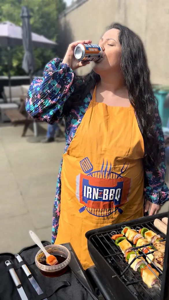 A person with long hair drinks from a can while wearing a yellow apron reading IRN-BBQ. They stand by a grill with skewers, and theres a table with utensils and a bowl of sauce nearby. An outdoor setting with chairs and an umbrella is in the background.