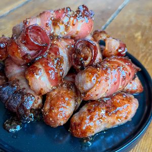 A close-up of glazed bacon-wrapped sausages piled on a black plate. The glossy coating highlights the crispy texture of the bacon, and the sausages have a rich, savory appearance. The wooden tabletop is visible in the background.