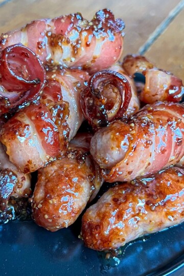 A close-up of glazed bacon-wrapped sausages piled on a black plate. The glossy coating highlights the crispy texture of the bacon, and the sausages have a rich, savory appearance. The wooden tabletop is visible in the background.
