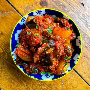 A colorful bowl of ratatouille with chunky pieces of eggplant, tomatoes, bell peppers, and herbs, placed on a wooden table. The dish is vibrant with rich red and green hues. The bowl has a blue and white floral pattern around its rim.