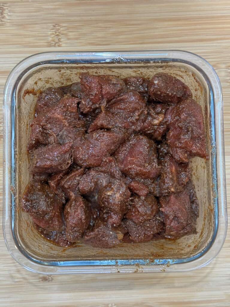 Chunks of raw meat coated in dark red marinade sit in a square glass container on a wooden surface.