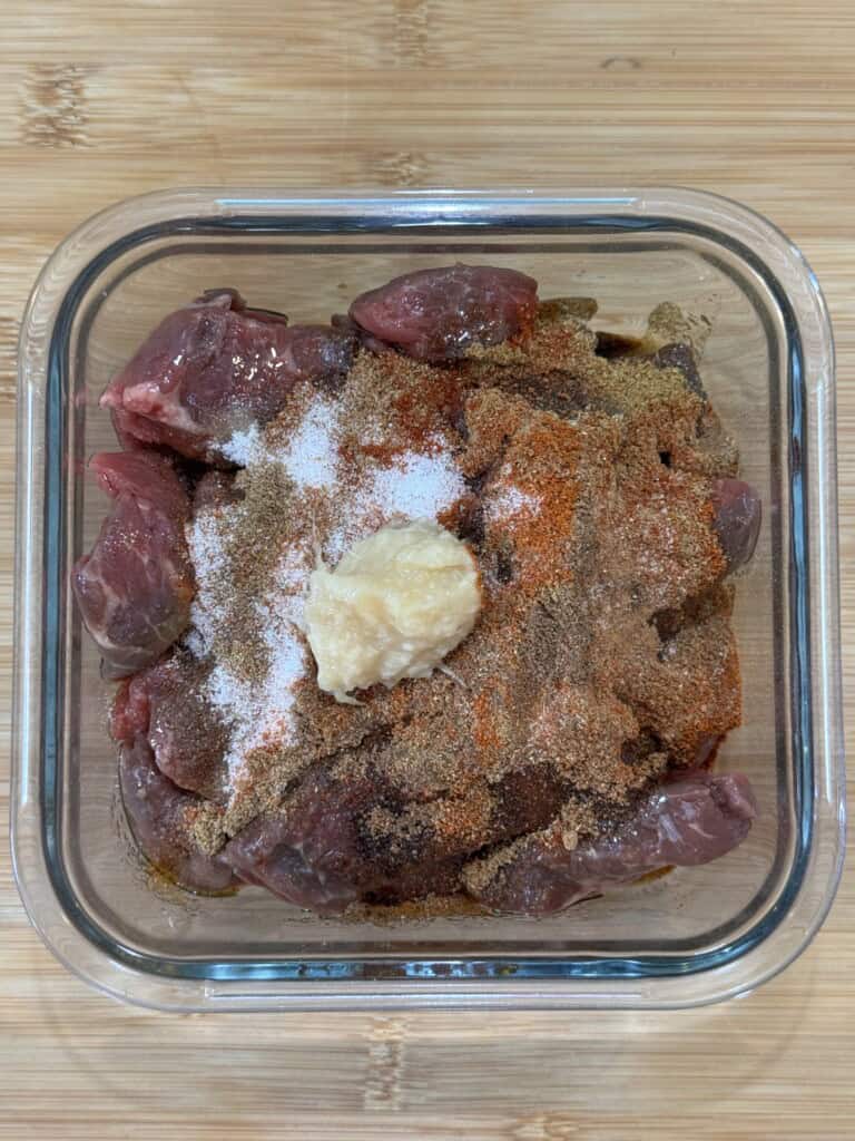 Raw chunks of meat in a glass container topped with various spices and a dollop of minced garlic, placed on a wooden surface.