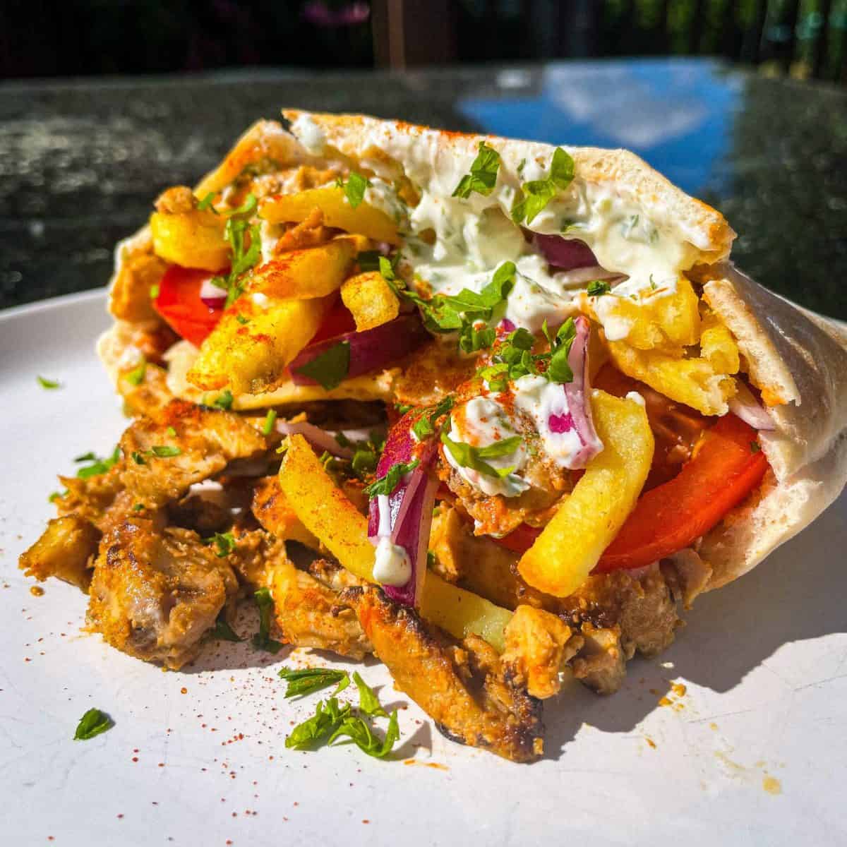 A close-up of a gyro with grilled meat, tomatoes, red onions, french fries, and tzatziki sauce wrapped in pita bread. Its garnished with chopped herbs and is served on a white plate in a sunlit setting.