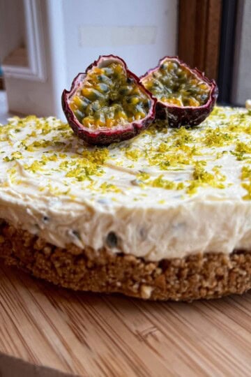 A cheesecake with a crumbly base and creamy topping sits on a wooden platter. It is garnished with lime zest and topped with halved passion fruits. The dessert is placed by a window, allowing natural light to highlight its texture.