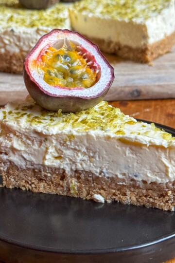A slice of creamy cheesecake topped with a halved passion fruit sits on a black plate. In the background, the rest of the cheesecake is visible on a wooden board. The cheesecake has a crumbly base and a yellowish topping.