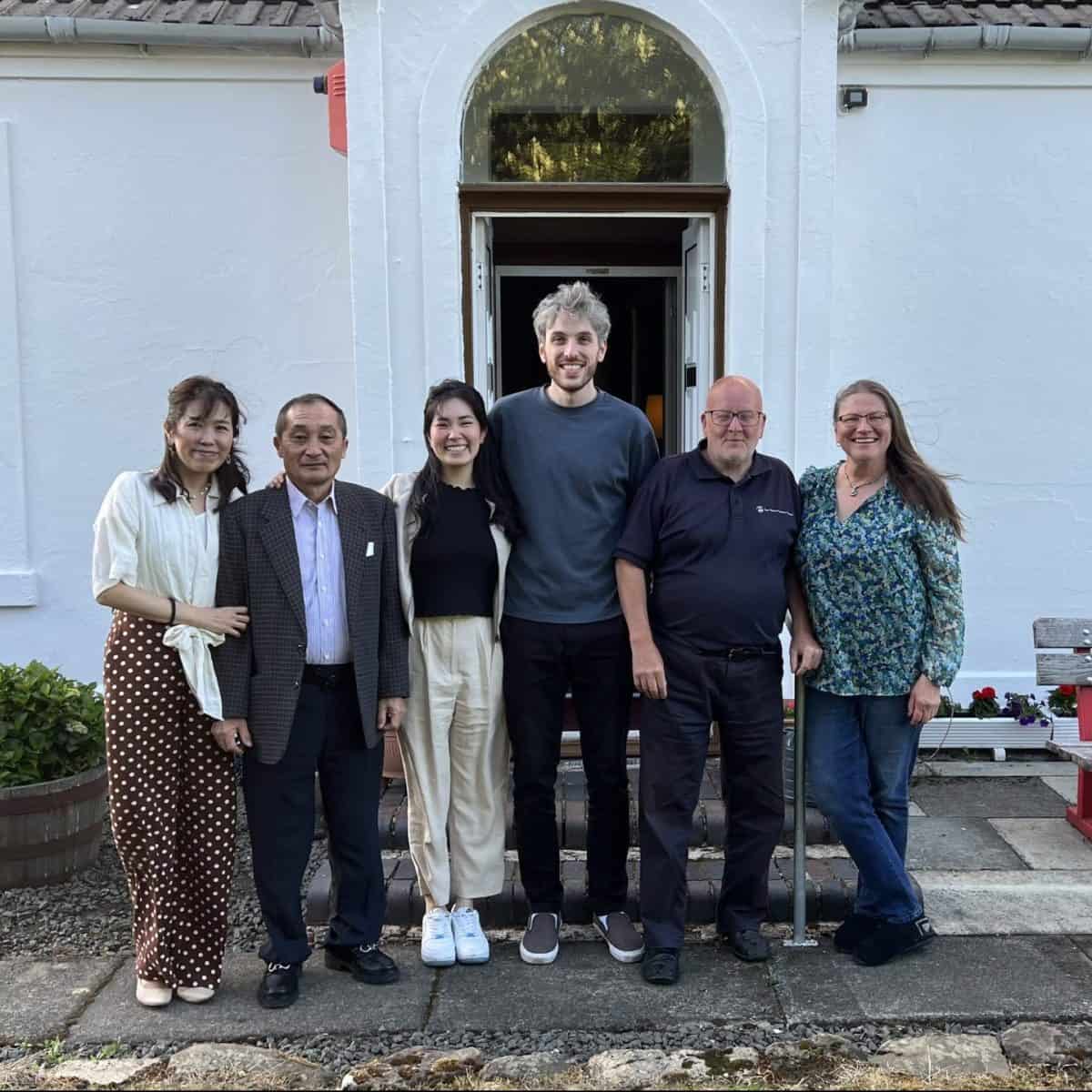 Six people stand smiling in front of a white building with an arched entrance. The group includes four adults of varied ages and two younger adults. They stand closely, with arms around each other, against a backdrop of potted plants and a bench.