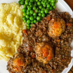 A plate of mashed potatoes, green peas, and a stew with meat and vegetables, topped with three golden-brown dumplings.