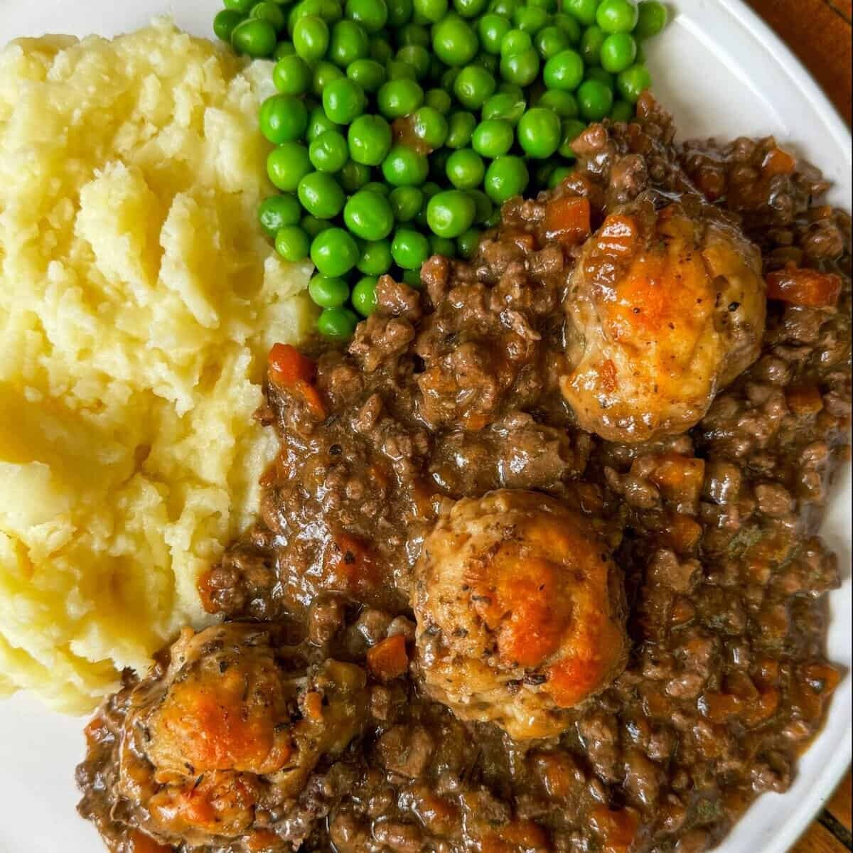 A plate of mince and dumplings: mashed potatoes, green peas, and a stew with meat and vegetables, topped with three golden-brown dumplings.