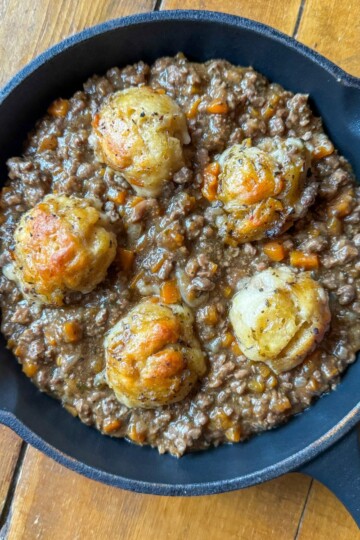 A black skillet on a wooden table contains a savory dish of minced meat mixed with carrots and herbs. Five golden-brown dumplings sit on top, partially embedded in the meat mixture.