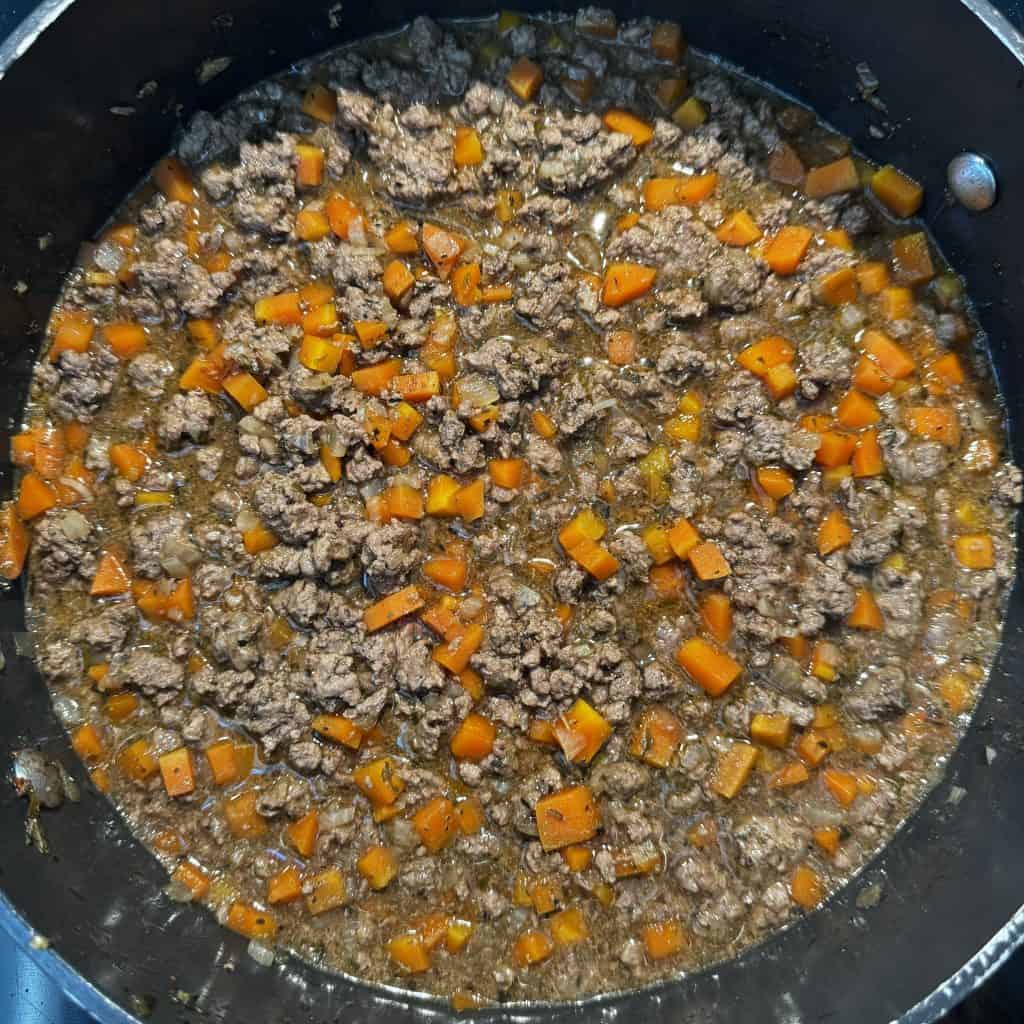 Mince and tatties: ground beef and diced carrots cooking in a black pan, with visible herbs and spices mixed in. The ingredients are simmering in a broth, giving a savory appearance.
