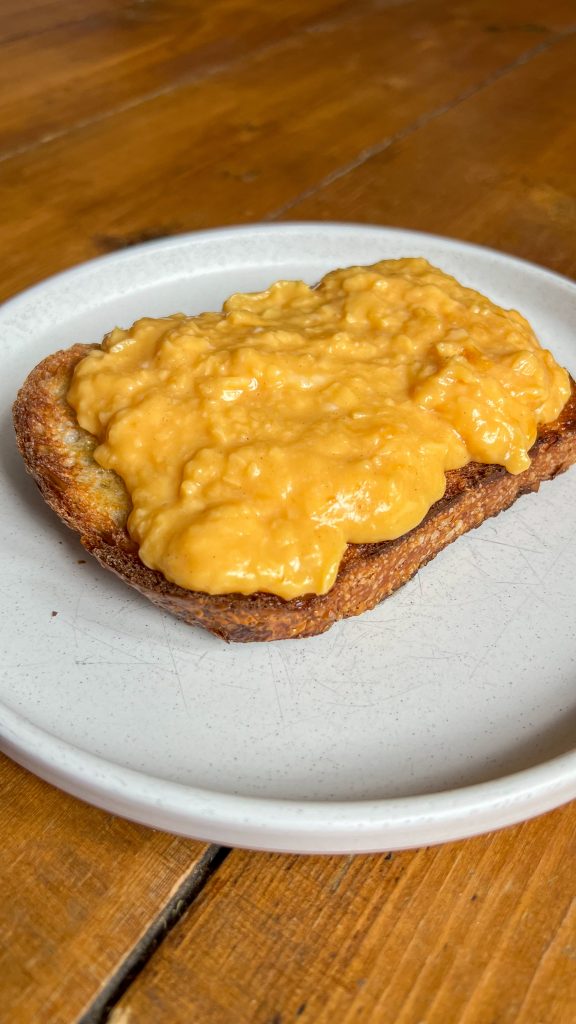 A slice of toasted bread topped with smooth scrambled eggs sits on a white plate. The eggs have a rich, creamy texture and a golden color. The plate is on a wooden table.