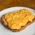 A slice of toasted bread topped with smooth scrambled eggs sits on a white plate. The eggs have a rich, creamy texture and a golden color. The plate is on a wooden table.