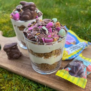 Two glasses of layered dessert with crushed candies and chocolate bunnies on top sit on a wooden board in a grassy area. A chocolate bunny is beside the glasses, and two colorful candy bags are nearby.