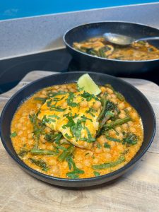 A black bowl filled with chickpeas, green beans, and spinach in a creamy orange sauce, topped with fresh cilantro and a lime wedge. A pan with more of the dish is blurred in the background. The bowl is placed on a wooden surface.