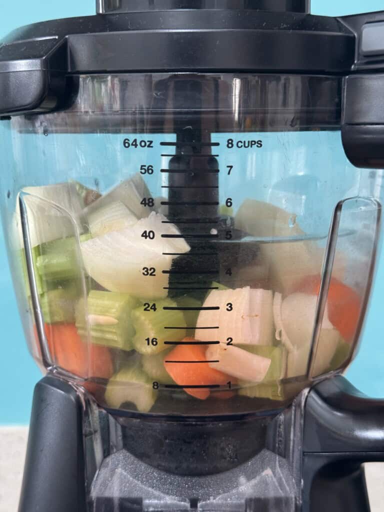 A close-up of a blender filled with chopped vegetables, including carrots, onions, and celery, with water up to the 32-ounce mark. Measurement markings are visible on the side.
