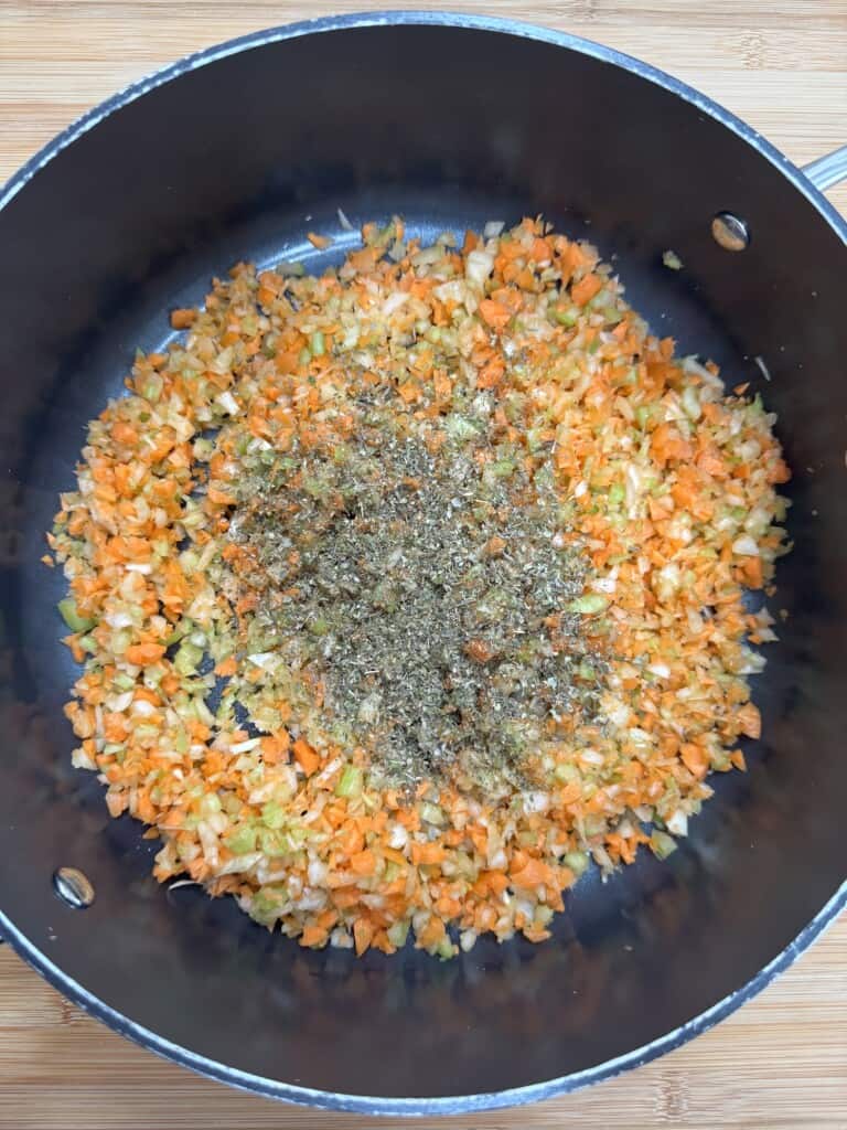 Chopped carrots, celery, and onions sauté in a large skillet, with a heap of dried herbs and spices added on top, ready to be mixed together.