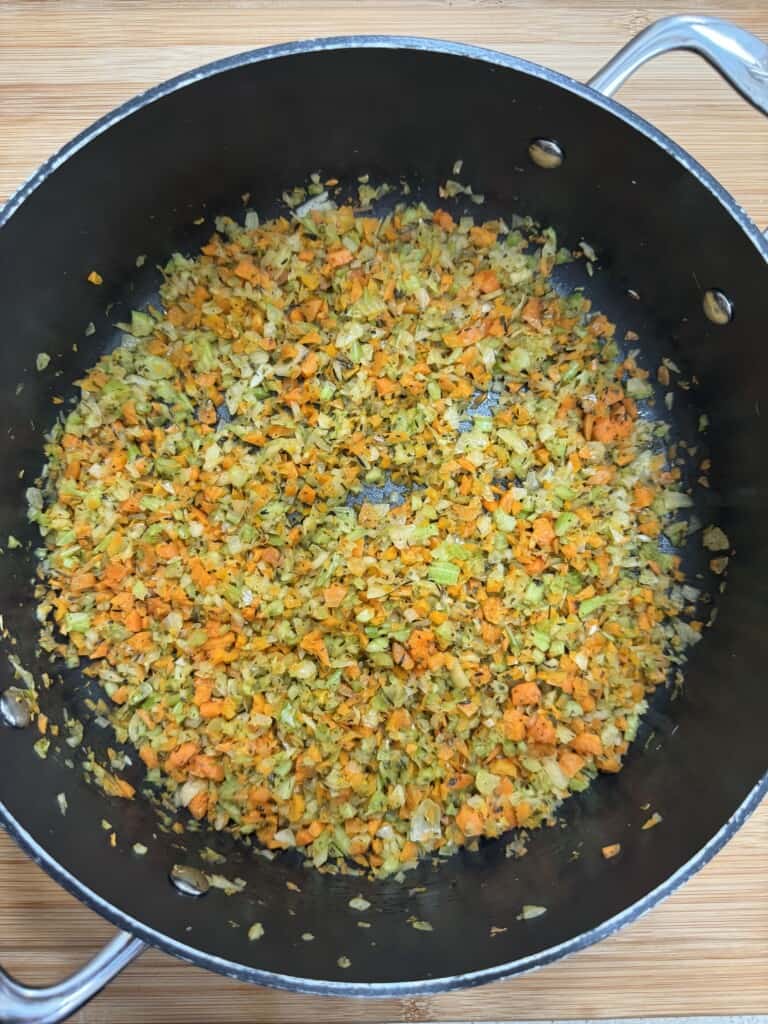 Finely chopped carrots, onions, and celery sautéing in a large black pot with silver handles, set on a wooden surface. The vegetables are evenly mixed and appear lightly cooked.