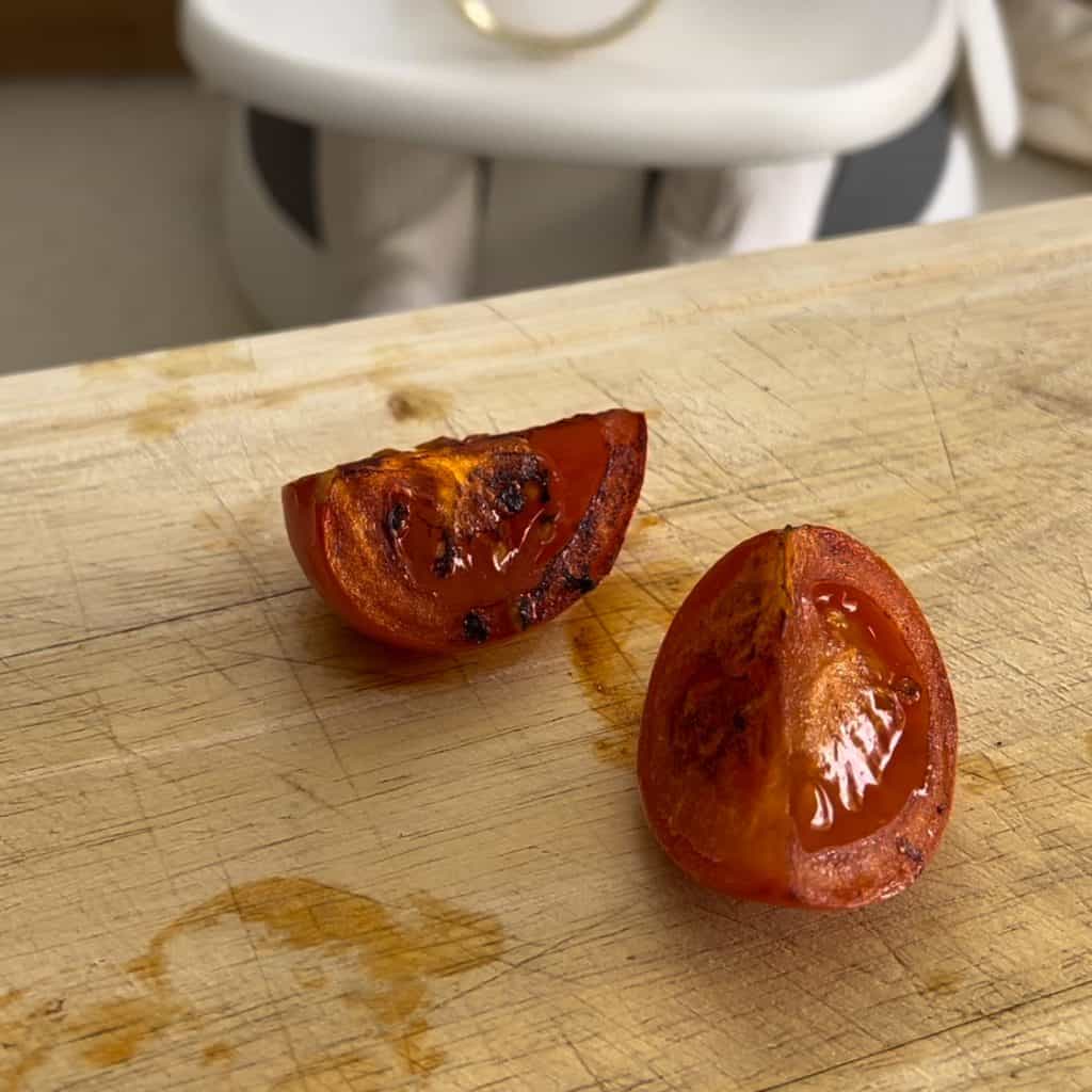 Two grilled tomato halves rest on a wooden cutting board. The tomatoes appear slightly charred, showing a juicy interior. The background features a blurred white and gray object.