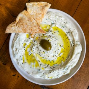 A bowl of creamy tzatziki topped with olive oil, a single olive, and sprinkled with herbs. Two pita bread triangles are placed on the side. The dish is on a wooden table.