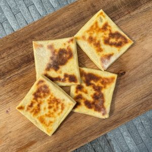 Four golden-brown, square-shaped flatbreads lie on a wooden board. They have a slightly crispy, browned surface, suggesting they are freshly cooked. The background features a textured, gray surface.