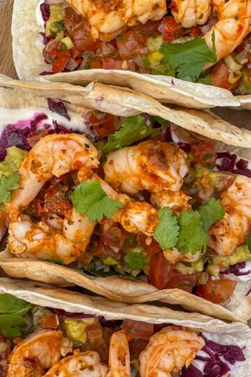 Three shrimp tacos on a wooden board, filled with spicy shrimp, fresh tomatoes, shredded red cabbage, cilantro, and a dollop of sour cream. The tacos are arranged closely, showcasing their colorful toppings.