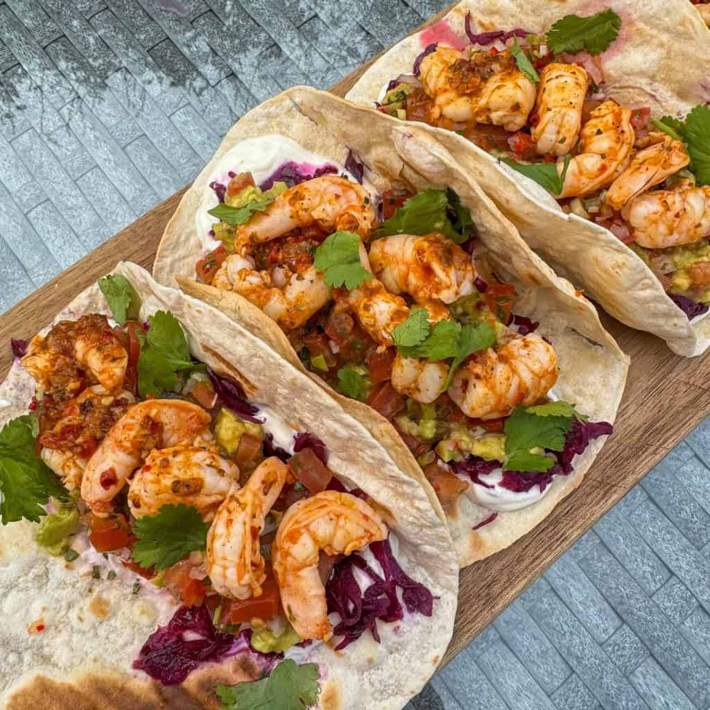 Three soft tacos with grilled shrimp, cabbage, diced tomatoes, creamy sauce, and cilantro are arranged on a wooden board. The tacos are placed on a textured gray surface.