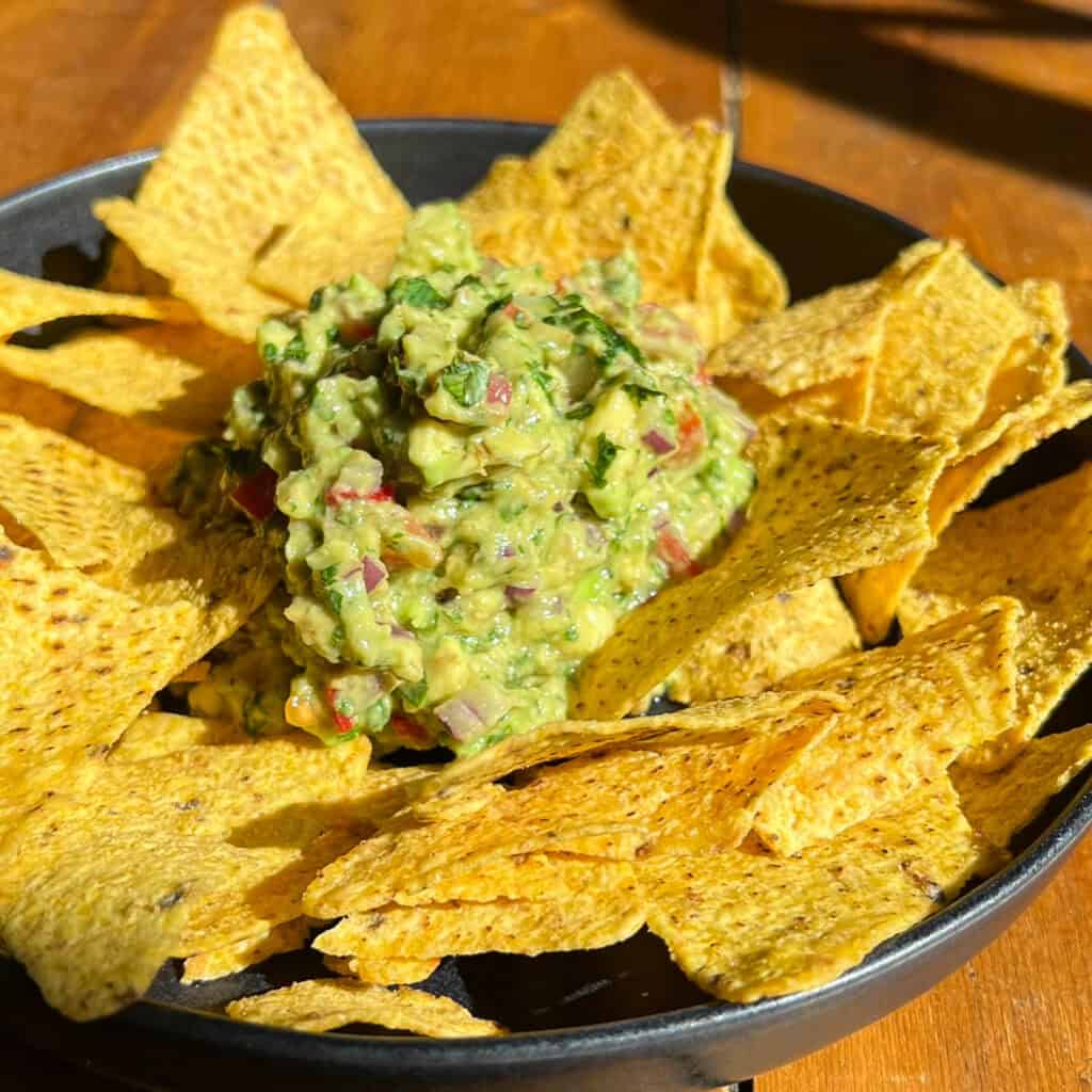 A black bowl filled with triangular corn tortilla chips surrounds a mound of chunky guacamole. The guacamole is chunky, with visible bits of red onion, cilantro, and tomato, set against a wooden table. The image is brightly lit by natural light.