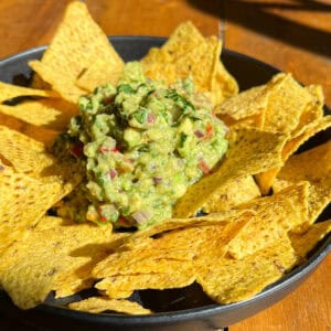 A black bowl filled with triangular corn tortilla chips surrounds a mound of chunky guacamole. The guacamole is chunky, with visible bits of red onion, cilantro, and tomato, set against a wooden table. The image is brightly lit by natural light.