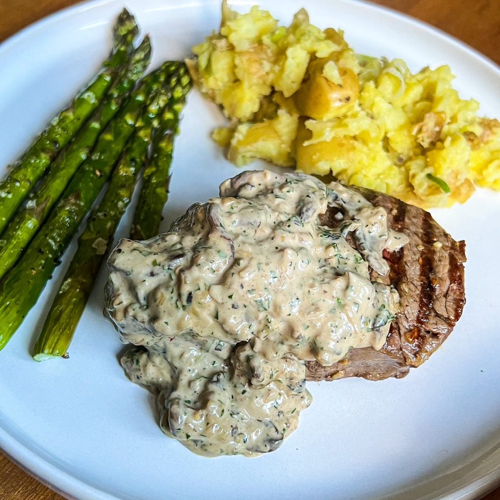 A plate with grilled steak covered in creamy mushroom sauce, served alongside roasted asparagus and a portion of mashed potatoes with bits of skin.