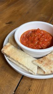 A white bowl filled with red pepper spread sits on a plate. Surrounding the bowl are slices of pita bread, arranged neatly on a wooden table.