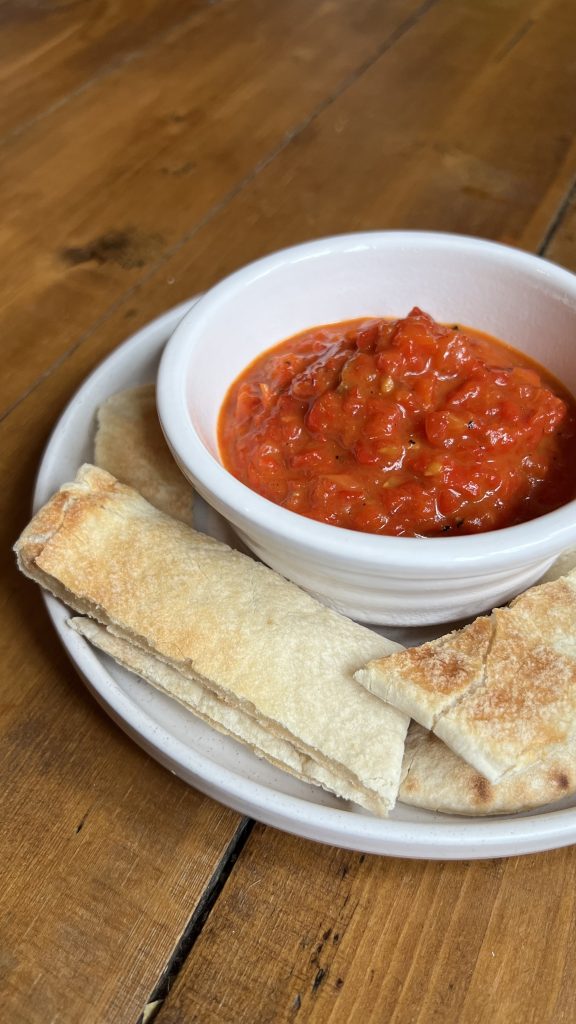 A white bowl filled with red pepper spread sits on a plate. Surrounding the bowl are slices of pita bread, arranged neatly on a wooden table.