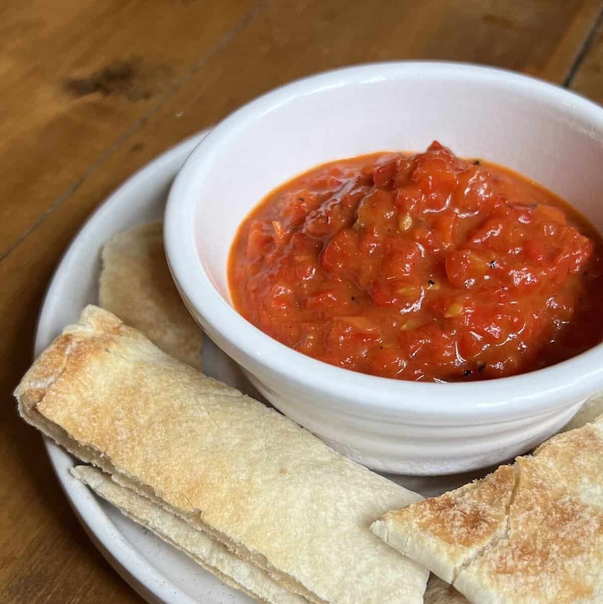 A white bowl filled with red pepper dip sits on a plate. Surrounding the bowl are slices of pita bread, arranged neatly on a wooden table.