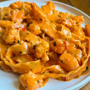 A close-up of a plate filled with creamy pasta topped with pieces of chicken or seafood, garnished with herbs. The sauce appears rich and orange-colored, coating the wavy pasta evenly.