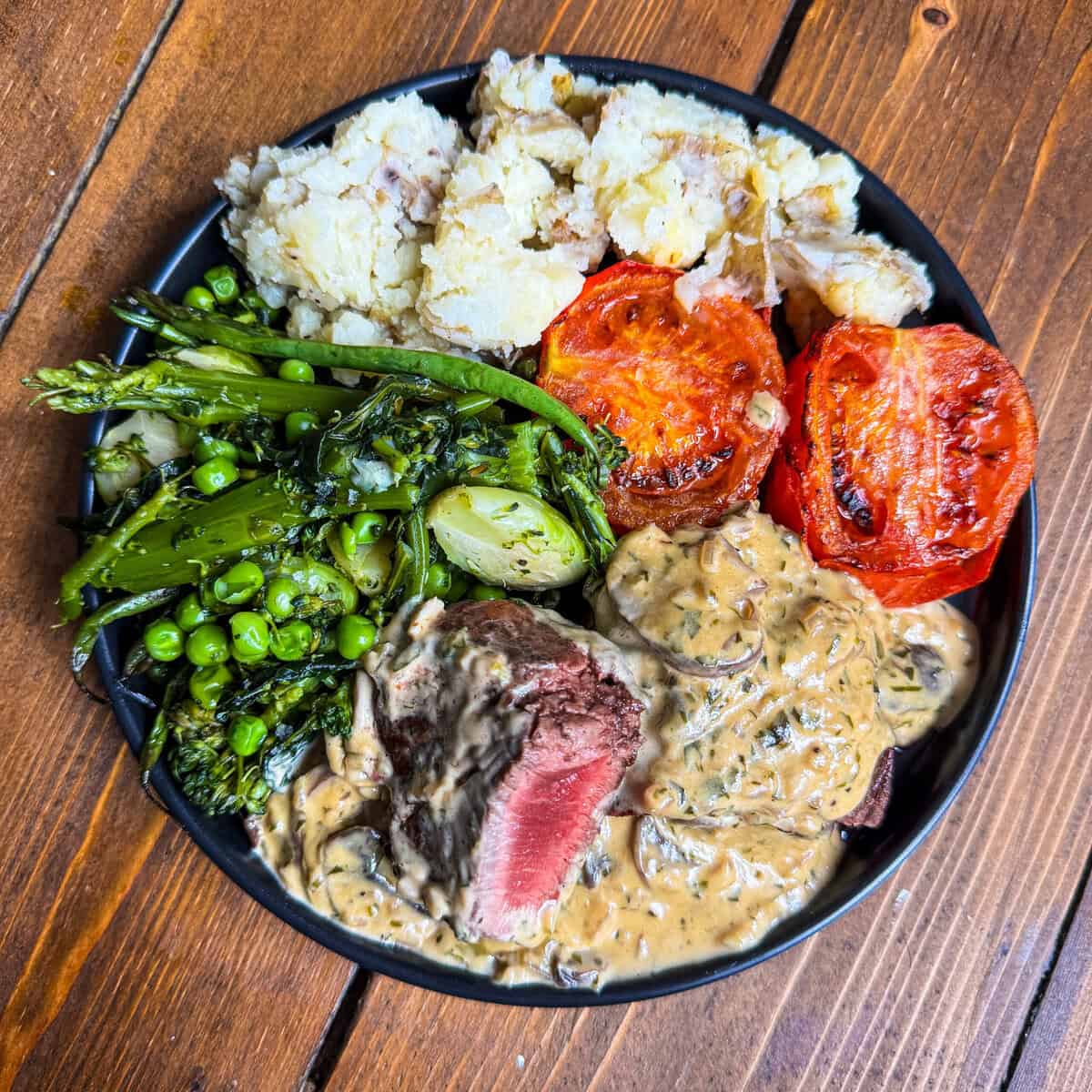 A plate with mashed potatoes, roasted tomatoes, sautéed greens and peas, and sliced beef topped with creamy Diane Sauce with mushrooms, served on a wooden table.