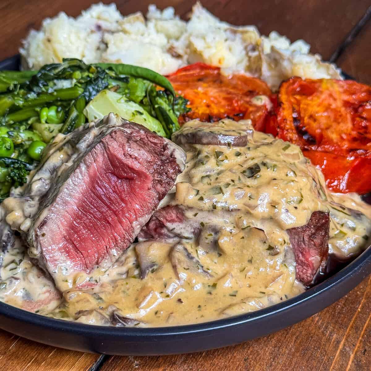 A plate of steak with creamy Diane sauce with mushrooms, mashed potatoes, roasted tomatoes, and sautéed broccolini, served on a wooden table. The steak is sliced to show a rare, red center.