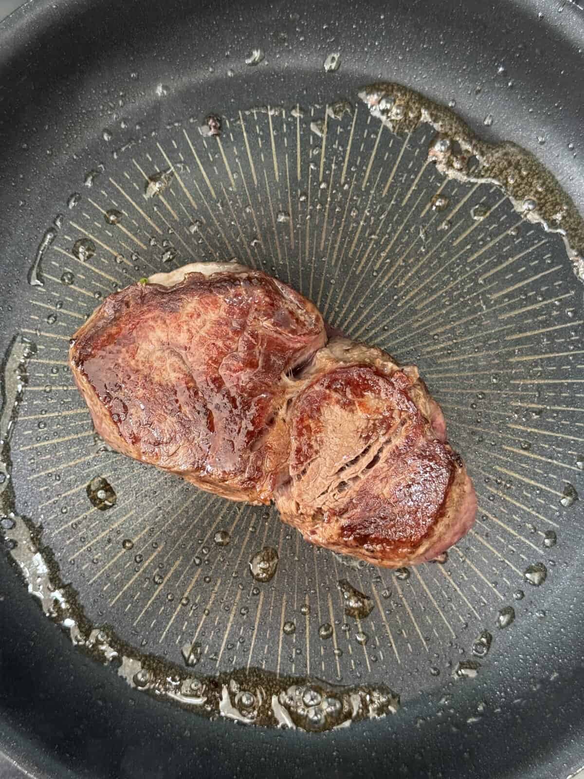 A seared steak with a browned crust is cooking in a black frying pan with a small amount of oil around it.