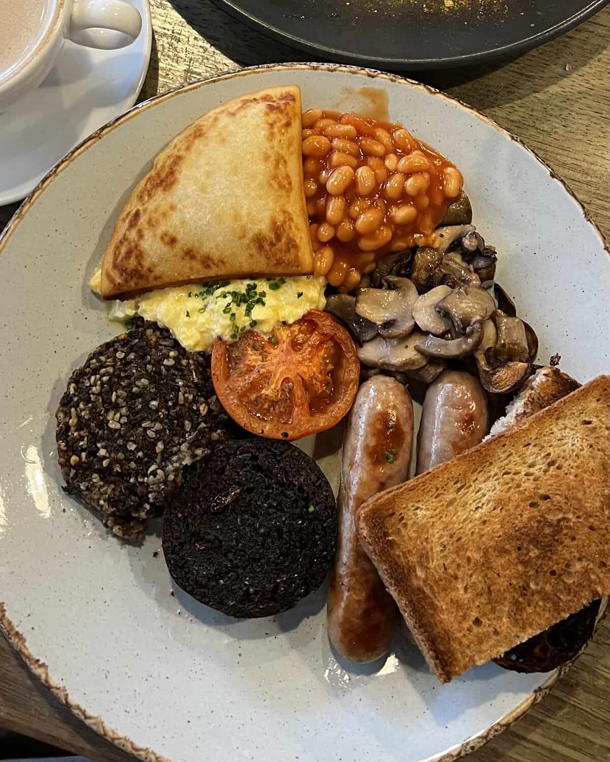 A plate with a full Scottish breakfast including sausages, bacon, black pudding, grilled tomato, toast, scrambled egg, baked beans, mushrooms, potato scone, and a cup of coffee on the side.