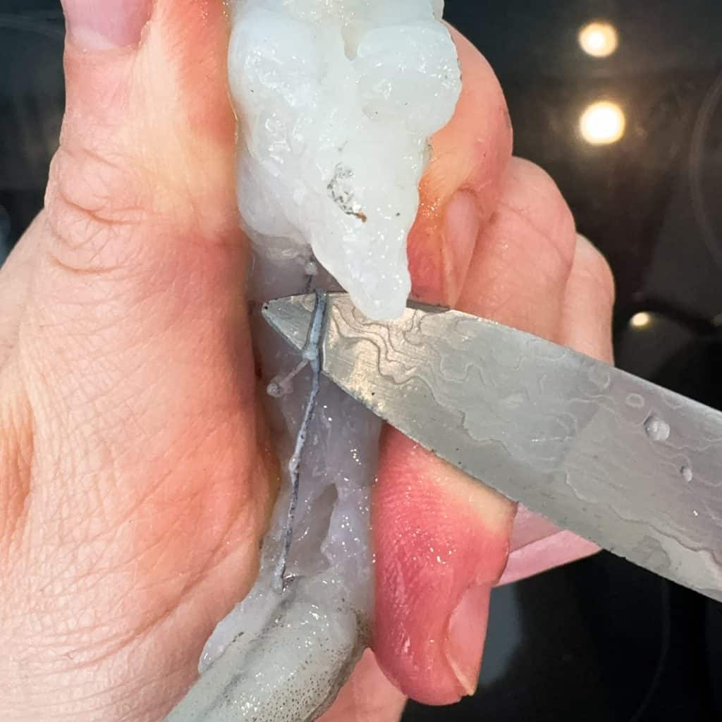 A hand holding a raw shrimp while cutting it with a knife along the back to remove the vein. The shrimp appears translucent and slightly curved, with some water droplets on its surface. The background shows a dark kitchen counter.