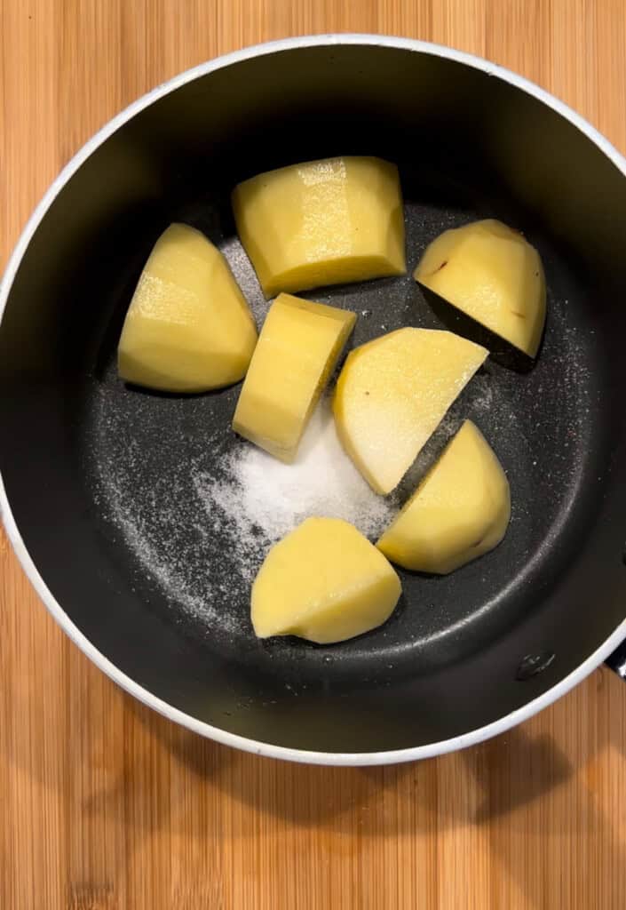 A black saucepan containing peeled and chopped raw potatoes with a sprinkle of salt, placed on a wooden surface.