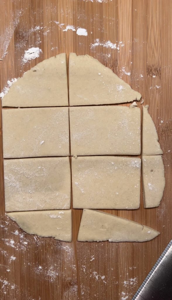Rectangular pieces of dough are cut on a floured wooden surface, with a few irregular scraps along the edge and a knife partially visible in the bottom right corner.