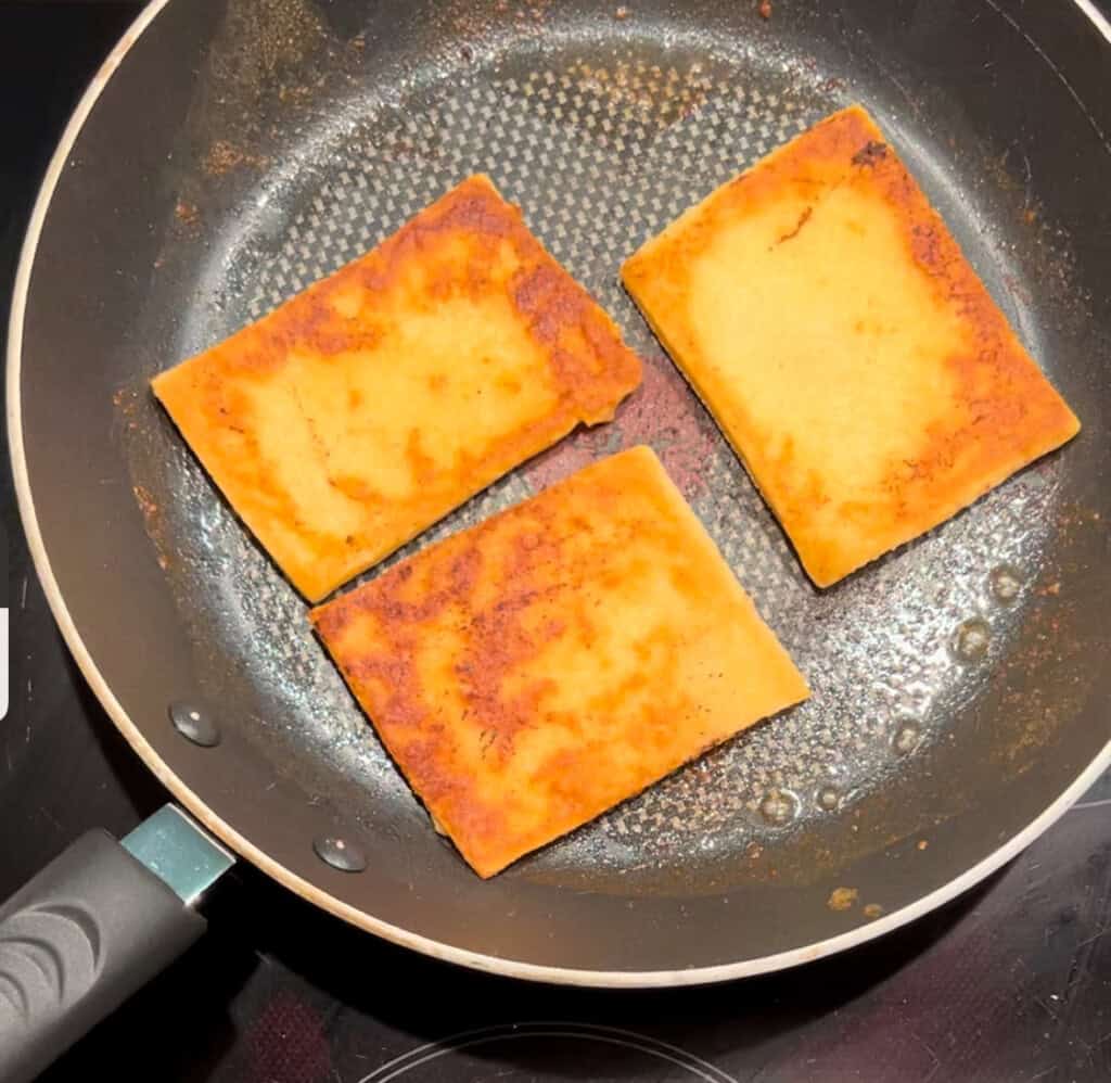Three golden-brown rectangular tattie scones are being fried in a non-stick frying pan. The pieces have crispy, browned surfaces and are evenly spaced in the pan.