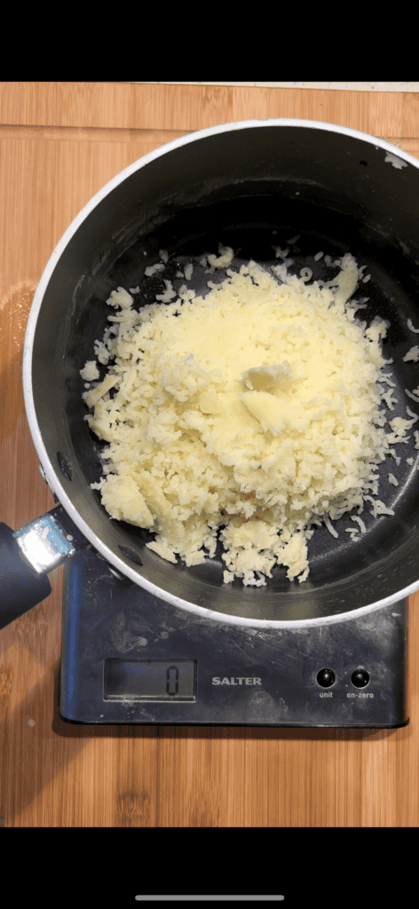 A small pan with grated cheese sits on an electric kitchen scale, which reads nought. The pan is placed on a bamboo surface.