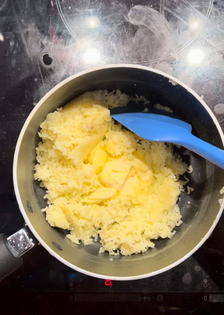 A saucepan on a hob contains mashed potatoes with a large knob of butter on top and a blue spatula resting inside the pan.